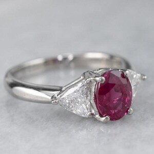 May include: A silver ring with a central oval ruby and two triangular diamonds. The ruby is a deep red hue, and the diamonds sparkle. The ring's band is polished silver, creating a sophisticated and timeless piece of jewellery.