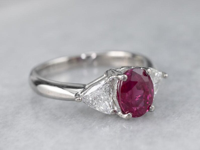 May include: A silver ring with a central oval ruby and two triangular diamonds. The ruby is a deep red hue, and the diamonds sparkle. The ring's band is polished silver, creating a sophisticated and timeless piece of jewellery.