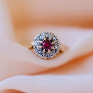 Op de afbeelding: Een gouden ring met een centrale rode edelsteen, omringd door kleinere heldere stenen. De zetting van de ring heeft een bloemmotief. De ring ligt op een zachte, perzikroze stof, wat een delicate en elegante uitstraling creëert.