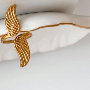 May include: A gold-colored ring with a wing design. The ring features two detailed wings, one on each side, with intricate feather detailing. The ring is set against a white background.