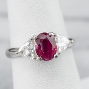 May include: A silver ring with a large, oval ruby gemstone, set between two triangular diamonds. The ruby is a deep red colour, and the diamonds are clear. The ring is displayed against a white background.