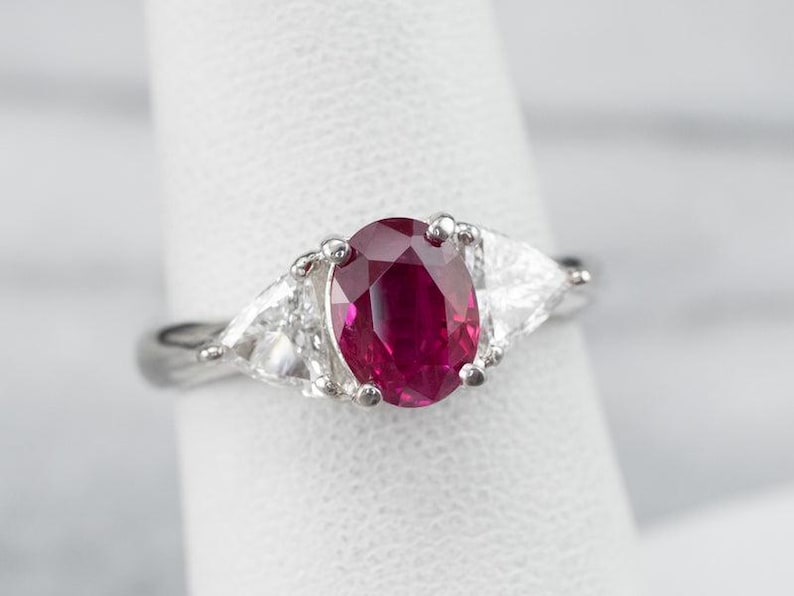 May include: A silver ring with a large, oval ruby gemstone, set between two triangular diamonds. The ruby is a deep red colour, and the diamonds are clear. The ring is displayed against a white background.