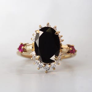 May include: A gold ring featuring a large, oval-cut black gemstone as the centerpiece. The ring is accented with smaller white and red gemstones, creating a striking contrast. The design is ornate and elegant.