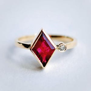 Op de afbeelding: Een gouden ring met een grote, levendige rode, ruitvormige edelsteen als centraal punt. Een kleine, ronde, heldere edelsteen is ernaast geplaatst. De ring heeft een eenvoudige, gepolijste gouden band.