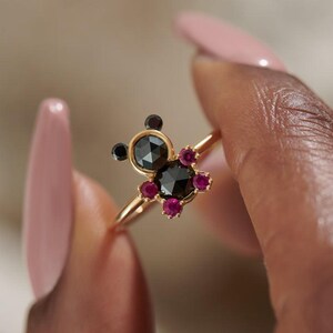 May include: A gold ring featuring a unique design with a central black gemstone surrounded by smaller, vibrant red gemstones. Two additional black gemstones are set on the ring's top, creating a distinctive, eye-catching piece of jewelry.