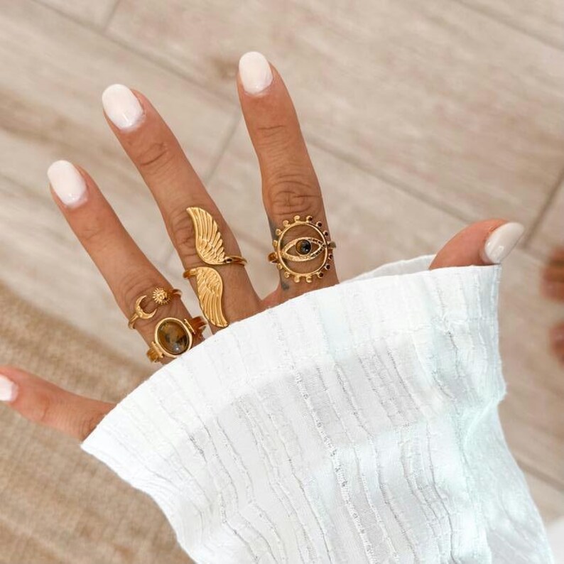 May include: A hand displaying five gold rings. The rings feature various designs, including wings, a sun and moon, and an eye. The rings are set against a white background.