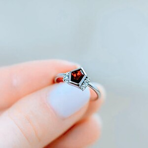 Puede incluir: Un anillo de plata con una gema roja oscura en forma de escudo geométrico en el centro. El anillo tiene pequeñas piedras de acento claras a cada lado de la piedra central. El anillo es sostenido por una persona.