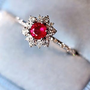 Op de afbeelding: Een delicate ring met een helderrode edelsteen in het midden, omringd door een halo van kleinere, sprankelende diamanten. De ring heeft een dunne band die is versierd met extra kleine diamanten, wat een stralend en elegant ontwerp creëert.