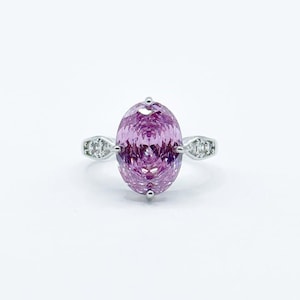 Puede incluir: Un anillo de plata con una gran piedra preciosa ovalada de color púrpura claro como pieza central. La banda está adornada con piedras preciosas más pequeñas y transparentes a cada lado de la piedra principal, creando un efecto brillante. El anillo está sobre un fondo blanco.