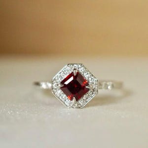 Op de afbeelding: Een zilveren ring met een vierkante, dieprode edelsteen in het midden, omringd door een halo van kleinere, heldere stenen. De band van de ring is ook versierd met kleine, heldere stenen, wat een sprankelend effect creëert. De ring is geplaatst tegen een zachte, neutrale achtergrond.