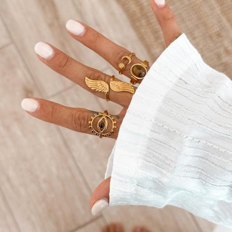 May include: A hand displays several gold-colored rings. One ring features a wing design, another has a sun and moon, and others have gemstone accents. The rings are worn on multiple fingers, showcasing a variety of styles and designs.