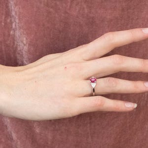 May include: A silver ring with a central oval-cut ruby, complemented by two triangular white gemstones. The ring is shown on a hand, set against a muted pink backdrop. The ruby is the focal point of the ring.