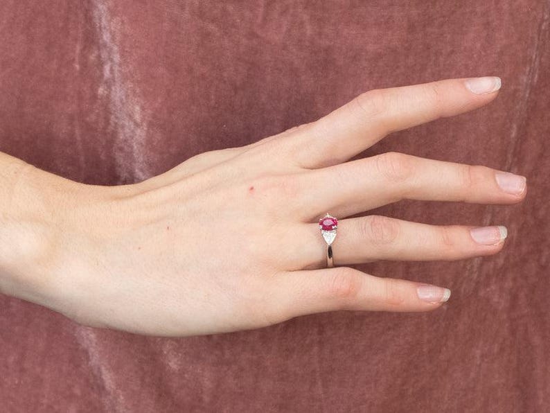 May include: A silver ring with a central oval-cut ruby, complemented by two triangular white gemstones. The ring is shown on a hand, set against a muted pink backdrop. The ruby is the focal point of the ring.