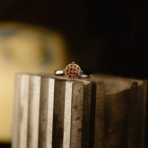 Può includere: Un anello d'oro con un design circolare, incastonato con piccole pietre preziose rosse. L'anello è posto su una superficie grigia strutturata, con uno sfondo sfocato. La fascia dell'anello è sottile e dorata.