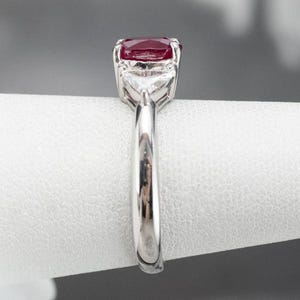 May include: A silver ring with a large, oval-cut, deep red gemstone as the focal point. Two smaller, triangular, clear gemstones are set either side of the main stone. The ring's band is polished silver, displaying a classic design.
