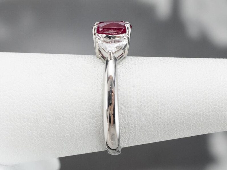 May include: A silver ring with a large, oval-cut, deep red gemstone as the focal point. Two smaller, triangular, clear gemstones are set either side of the main stone. The ring's band is polished silver, displaying a classic design.