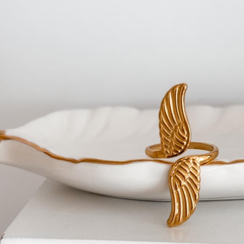 May include: A gold-colored ring with a wing design. The ring features two detailed wings that wrap around the finger. The ring is displayed on a white surface with a gold trim.