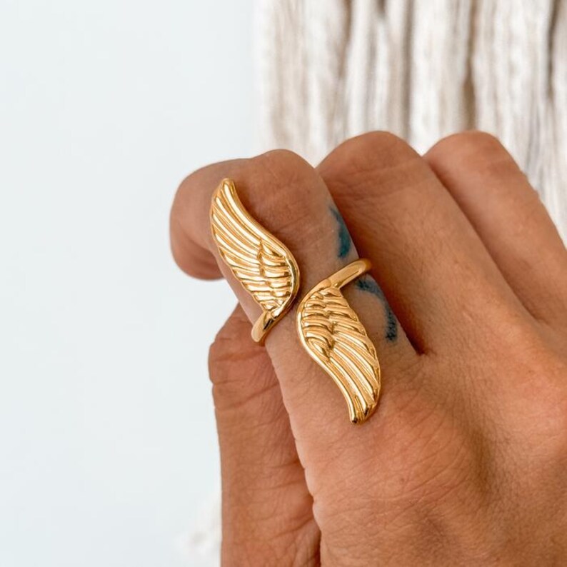 May include: A gold-colored ring with a wing design. The ring features two stylized wings, one on each side of the finger. The wings have detailed feather patterns. The ring is worn on a hand.