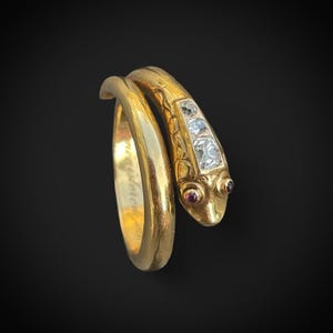 May include: A gold snake ring with a coiled design. The snake's head features three clear stones and two small red stones for eyes. The ring is set against a black background.