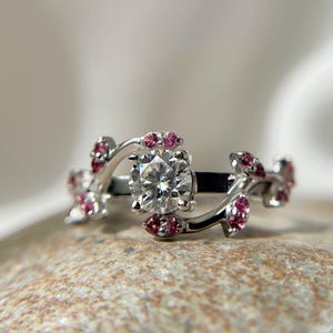 Op de afbeelding: Een zilveren ring met een grote, heldere, rond geslepen middensteen. De ring heeft een bloemmotief met kleine roze edelstenen. De band buigt om de middensteen en is versierd met de roze edelstenen, wat een delicate en elegante uitstraling creëert.