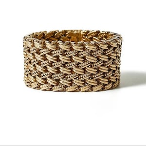 Może przedstawiać: Złota bransoletka o plecionym wzorze. Bransoletka jest prostokątna i posiada szereg przeplatanych ogniw, tworzących teksturowaną powierzchnię. Metal ma polerowane wykończenie, odbijające światło. Biżuteria jest umieszczona na białym tle.