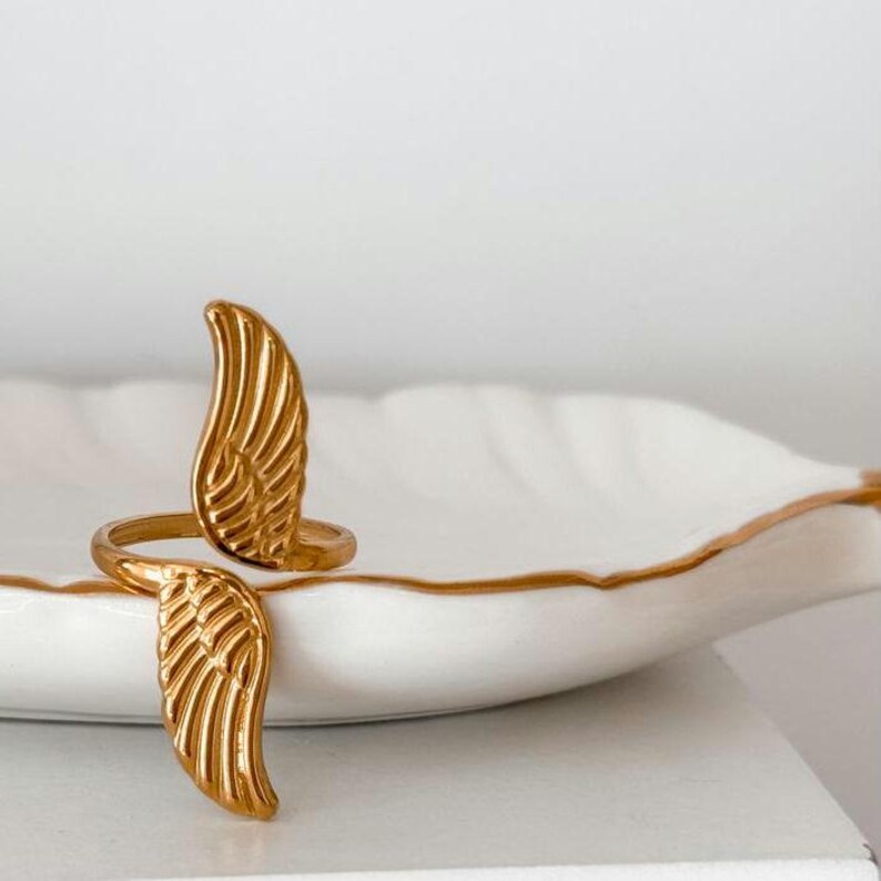 May include: A gold-colored ring with a wing design. The ring features two detailed wing shapes that wrap around the finger. The ring is displayed on a white, leaf-shaped dish with a gold trim.