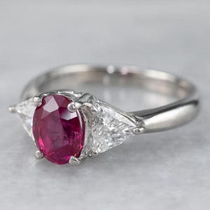 May include: A silver ring with a central oval ruby and two triangular diamonds. The ruby is a rich red hue, and the diamonds are clear. The ring's band is polished. The ring is a classic design, suitable for various occasions.
