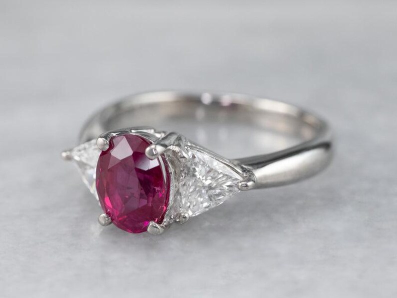 May include: A silver ring with a central oval ruby and two triangular diamonds. The ruby is a rich red hue, and the diamonds are clear. The ring's band is polished. The ring is a classic design, suitable for various occasions.