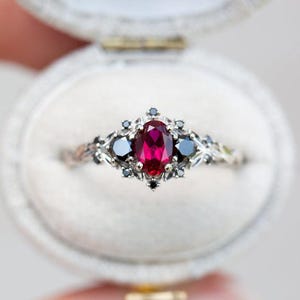 Op de afbeelding: Een zilveren ring met een ovale, dieprode edelsteen in het midden, geflankeerd door twee donkerblauwe stenen. De ring heeft ingewikkelde, bloemgeïnspireerde details en wordt gepresenteerd in een witte fluwelen ringdoos.