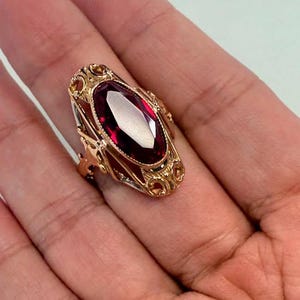 May include: A gold-toned ring featuring an oval, deep red gemstone. The ring has an ornate filigree design around the gemstone and on the band. The ring is held in a hand, showcasing its size and detail. A vintage-style piece.