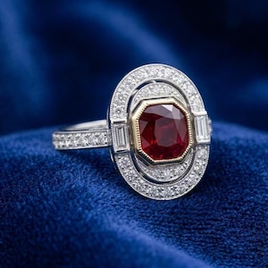 Peut inclure: Une bague ovale avec une grande pierre centrale en rubis facetté. Le rubis est entouré d'un halo de petits diamants étincelants. La monture de la bague est également ornée de diamants, créant un look luxueux et élégant. Posée sur un fond bleu velours.