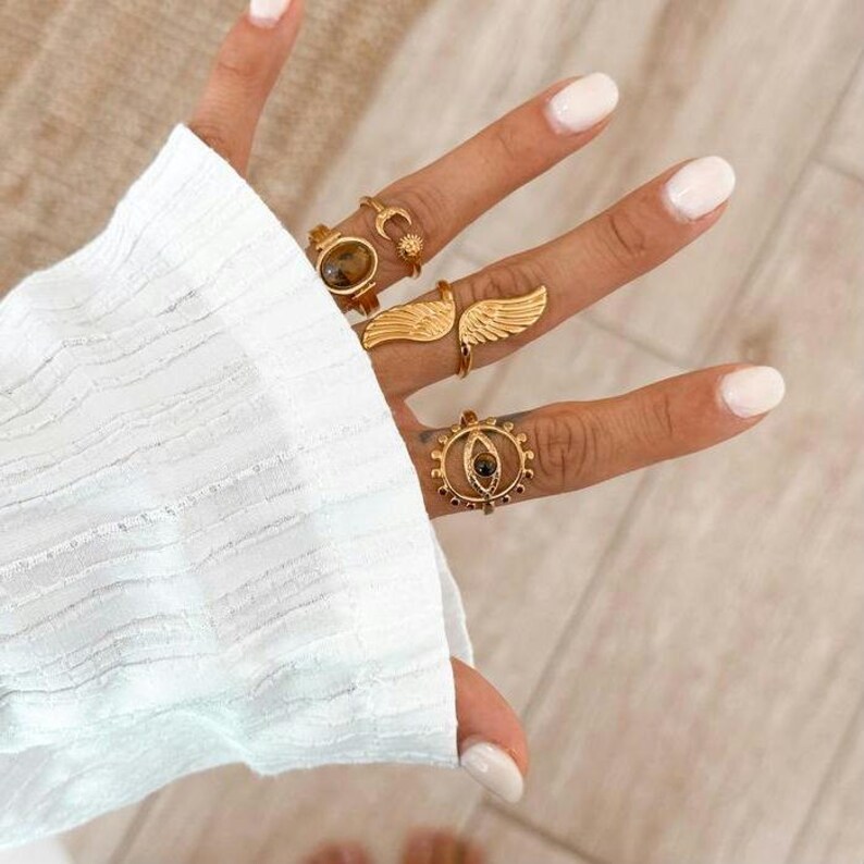May include: A hand displaying several gold-colored rings. The rings feature various designs, including a moon and sun, wings, and an eye-shaped design. The hand is wearing a white long-sleeved shirt.