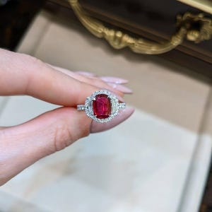 Op de afbeelding: Een schitterende ring met een levendige, rechthoekige rode edelsteen, omringd door een halo van kleinere, sprankelende witte diamanten. De ringband is ook versierd met diamanten, wat zorgt voor een luxueus en opvallend ontwerp.