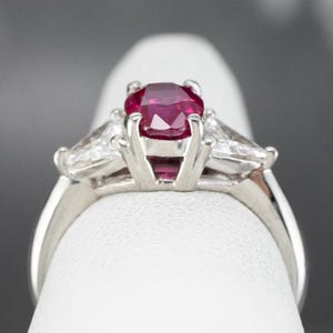 May include: A platinum ring with a large, oval-cut ruby as the central stone. Two smaller, triangular diamonds are set either side of the ruby. The ring's band is smooth and polished, highlighting the gemstones.