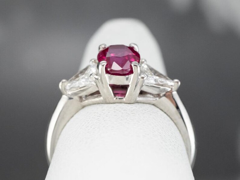 May include: A platinum ring with a large, oval-cut ruby as the central stone. Two smaller, triangular diamonds are set either side of the ruby. The ring's band is smooth and polished, highlighting the gemstones.
