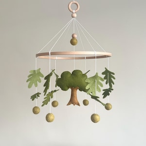 May include: A baby mobile featuring a wooden ring and a tree design. The mobile includes a felt tree, leaves, and round accents in shades of green and brown, suspended by white strings. The top has a wooden ring and a wooden bead.