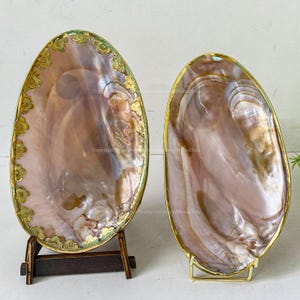 Peut inclure: Deux plats décoratifs ovales en forme de coquillage irisé. L'un est présenté sur un support en bois foncé, l'autre sur un support en métal doré. Les deux ont une bordure dorée. Les coquillages sont de couleur rose et marron nacré.