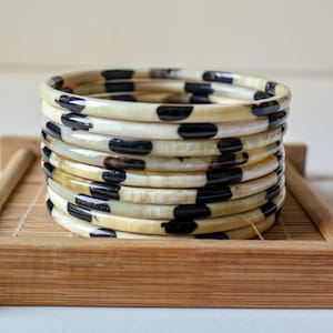 May include: A stack of ivory-colored bangles with black polka dot patterns. The bracelets are stacked on a small, rectangular wooden tray. The bangles have a glossy finish and a smooth texture. The overall aesthetic is simple and elegant.