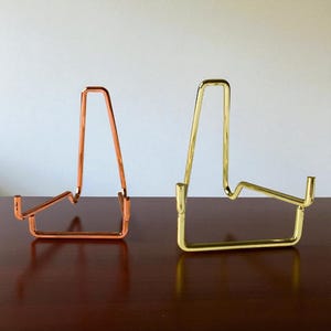 Peut inclure: Deux présentoirs métalliques. L'un est de couleur cuivre, l'autre est de couleur or. Les deux présentoirs ont un design similaire avec une base rectangulaire et un sommet arqué, adaptés pour tenir des assiettes ou des œuvres d'art.