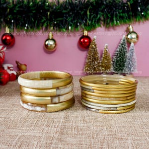 Peut inclure: Deux piles de bracelets dorés et blancs. L'une présente un motif marbré or et blanc, l'autre est principalement dorée. Les bracelets sont présentés sur une surface texturée avec des décorations de Noël en arrière-plan.