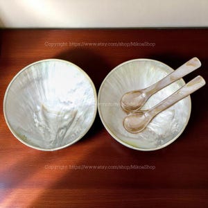 Puede incluir: Dos cuencos redondos de nácar iridiscente con un par de cucharas a juego. Los cuencos tienen un brillo nacarado con un borde de color claro. Las cucharas son de color marrón claro y tienen un acabado nacarado similar. Los cuencos y las cucharas están sobre una superficie marrón oscuro.