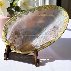Peut inclure: Un plat décoratif ovale en coquillage irisé, bordé d'or et orné de motifs floraux. Le plat est présenté sur un petit support en bois. La surface de la coquille présente des reflets nacrés roses, bleus et verts.