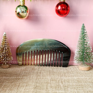 Puede incluir: Un peine de color marrón oscuro y marrón claro hecho de cuerno con una parte superior redondeada y 12 dientes. El peine está sobre una superficie de arpillera marrón. Dos pequeños árboles de Navidad artificiales y dos adornos navideños están en el fondo.