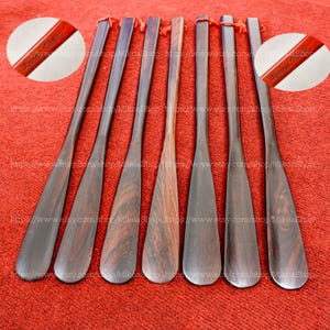 May include: Eight wooden shoe horns with a dark brown wood grain. The shoe horns are all the same size and shape, and they have a rounded tip. The shoe horns are arranged in a row on a red surface. The text "Tommy Tribe" is engraved on the top of each shoe horn.
