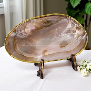 Peut inclure: Un plat décoratif ovale en forme de coquillage avec un bord doré, posé sur un support en bois foncé. L'intérieur de la coquille présente des tons irisés roses, bruns et crème. Le plat est présenté sur une surface blanche.