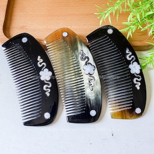Könnte beinhalten: Drei dekorative Haarkämme in Schwarz, Braun und Beige. Jeder Kamm hat ein geschwungenes Design mit einem weißen Blumen- und Wirbelmuster. Die Kämme haben eine abgerundete Oberseite mit einem kleinen, weißen kreisförmigen Akzent und feinen Zähnen.