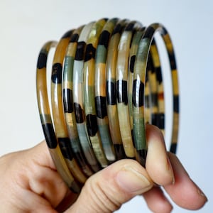 Puede incluir: Un conjunto de pulseras translúcidas con un diseño de carey. Las pulseras presentan una mezcla de colores, incluyendo negro, marrón y crema. Las pulseras se sostienen en una mano sobre un fondo blanco.