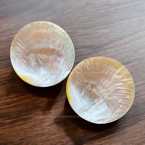 Puede incluir: Dos cuencos redondos de nácar blanco con un ligero brillo iridiscente. Los cuencos están sobre una superficie de madera marrón oscura.