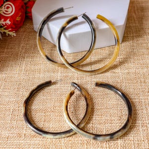 Peut inclure: Trois paires de grandes boucles d'oreilles en forme de cerceau en corne brune et noire. Les boucles d'oreilles sont sur un fond en tissu beige.