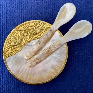 Peut inclure: Deux cuillères en nacre avec des accents dorés sur un plat en nacre avec un bord doré et un motif de poisson doré.